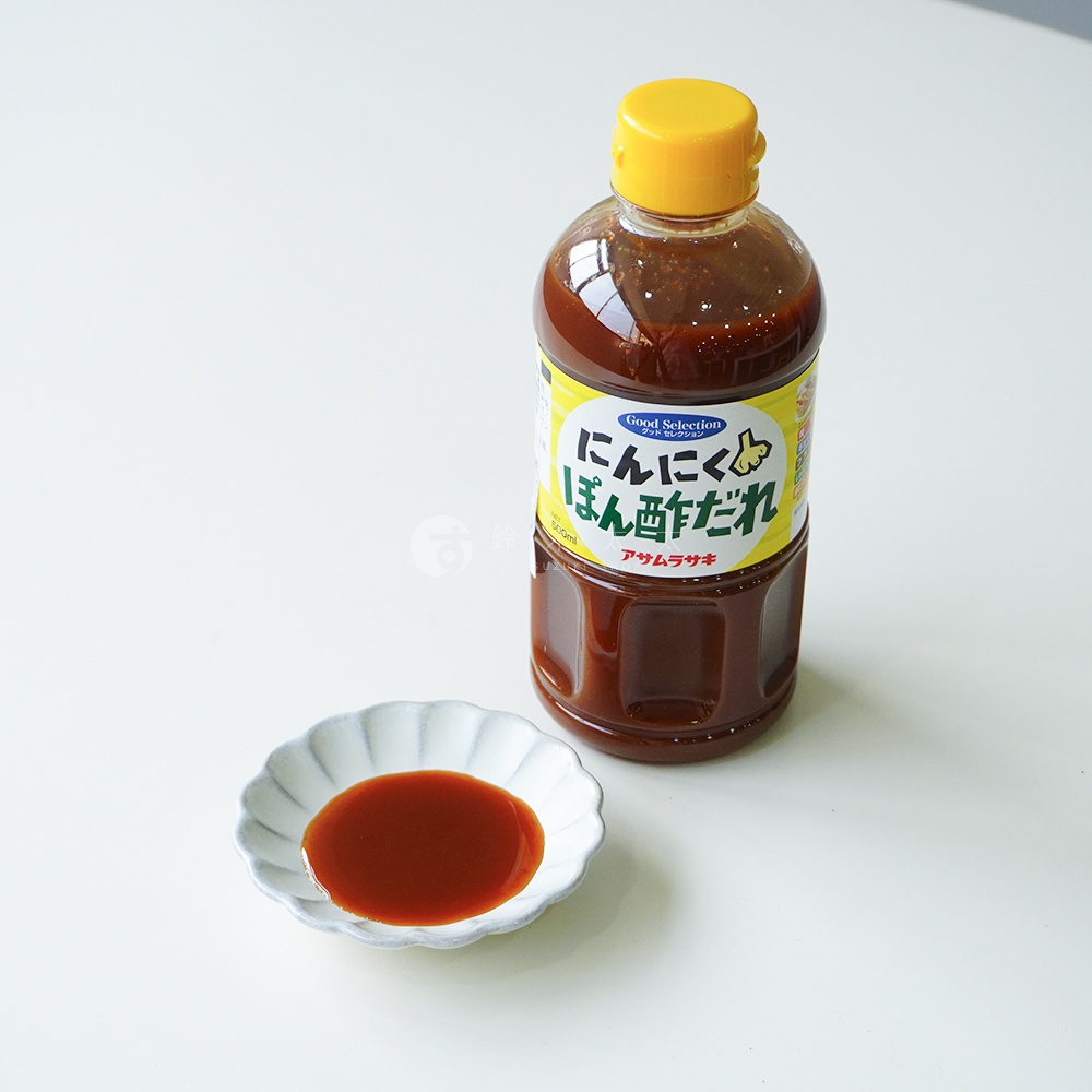 一瓶帶有黃色瓶蓋、裝在塑膠瓶中的深褐色液體調味醬，旁邊放著一個裝有少量相同深色醬汁的小碟子，瓶身標籤上寫有日文朝紫大蒜酸橘醋風味調味醬字樣。