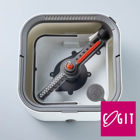 這是一款由上方視角拍攝的白色方形拖把清潔桶，內部設有旋轉清潔裝置，包含一條帶有白色刷毛與橘色組件的攪拌軸，桶身右下角有一處帶有水龍頭圖示的排水孔設計，右下角則標示著粉紅色的COGIT品牌商標。