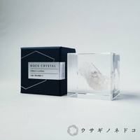 一個透明的壓克力立方塊，內部封存著一塊白色、半透明的水晶簇，水晶表面帶有細微的紋理和雜質。立方塊旁有一個深藍色的紙盒，盒蓋上印有銀色的標籤，標籤上寫有「ROCK CRYSTAL」和日文文字。
