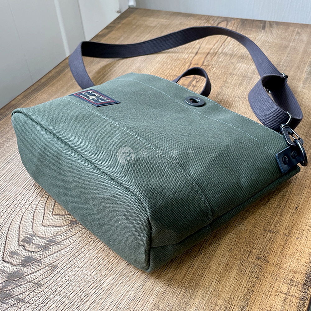 綠色純綿帆布迷你側背包，配有深灰色織帶肩帶，包身側面有精緻的縫線，正面有一個紅色的品牌標籤和一個金屬按鈕。