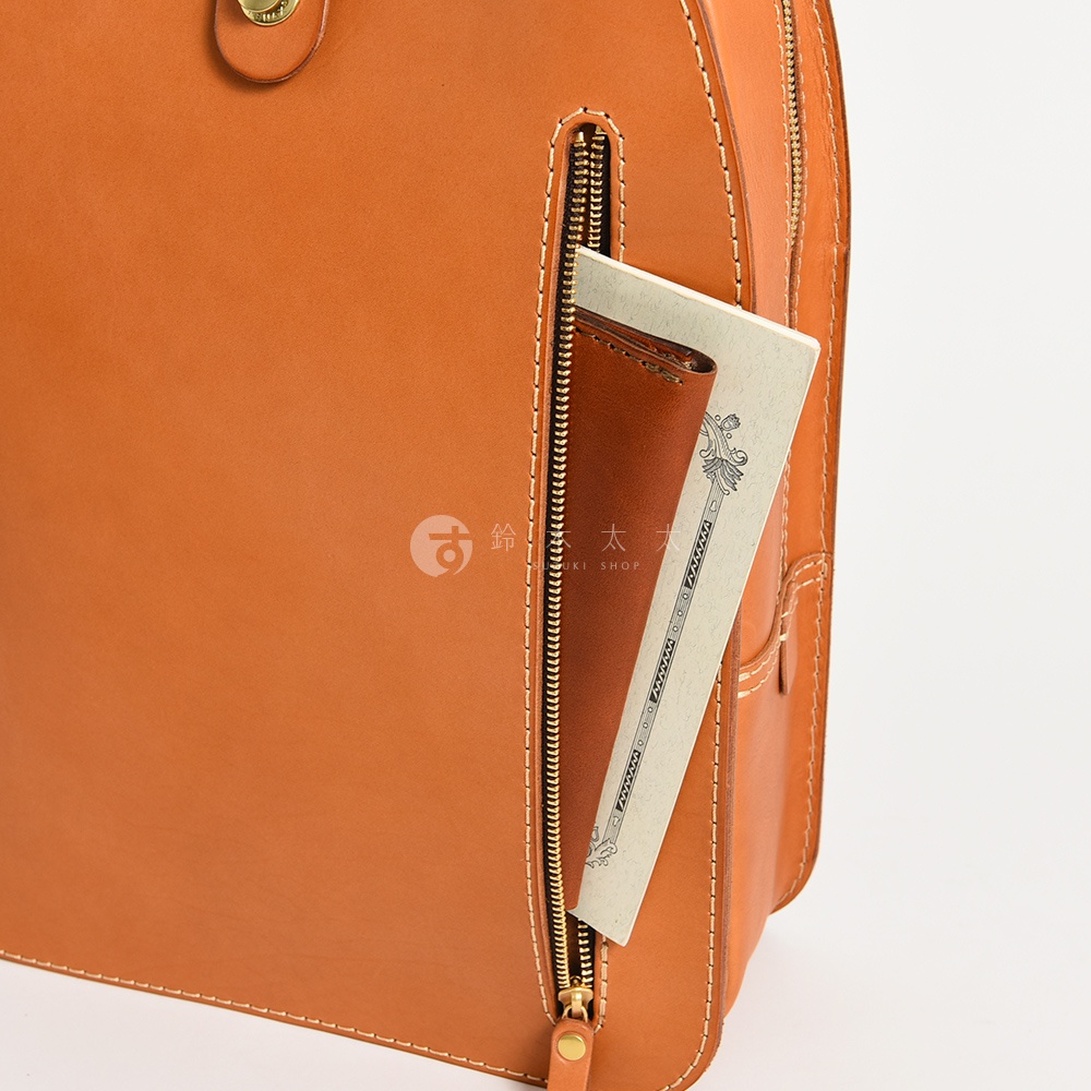 橘棕色皮革製成的兩用後背包，側面拉鍊開口處露出一個同色系皮革證件夾和一本淺米色筆記本。