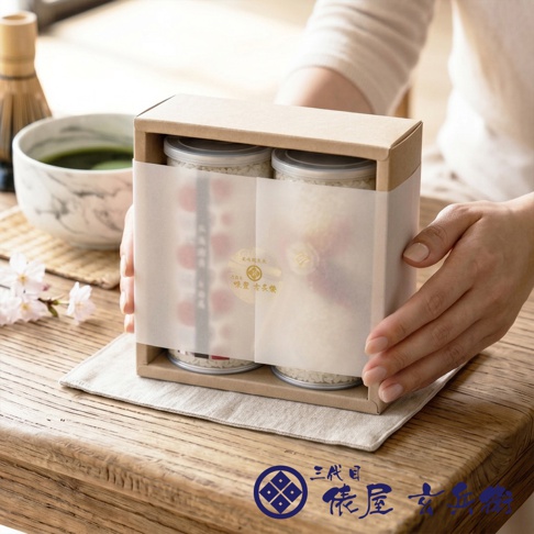 一位穿著白色上衣的人雙手輕握著三代目俵屋玄兵衛的北海道星月米禮盒，禮盒內裝有兩罐透明容器裝的米，包裝外層裹著半透明紙並貼有精緻圓形貼紙。桌面上擺放著盛有抹茶的茶碗、茶筅以及櫻花裝飾，整體呈現出日式雅致的氛圍。