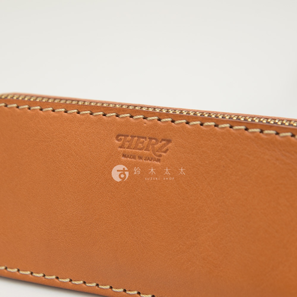 職人手作皮革三角弧形收納筆袋，淺棕色皮革，壓印有「HERZ」品牌標誌、「MADE IN JAPAN」字樣及日文「鈴木太太」與「SUZUKI SHOP」標誌，搭配金色拉鍊與米色縫線。
