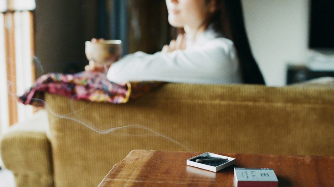 木質桌面上擺放著一盒白色的香氛火柴，內含一根燃燒的火柴，冒出細微的煙霧。旁邊另有一盒紅白色紙盒包裝的火柴。背景為黃色絨布沙發，上面搭著一塊色彩鮮豔的布料，一位女士的側臉模糊可見。