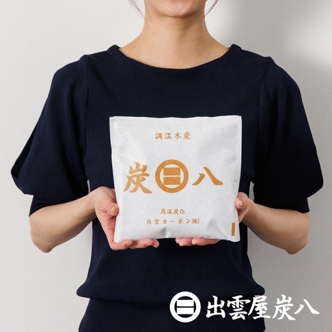 一位穿著深藍色上衣的女性，雙手捧著一包白色的出雲屋炭八小型調濕木炭，包裝袋中央印有金色的炭八字樣與品牌標誌，下方則有出雲屋炭八的品牌文字與圖標。