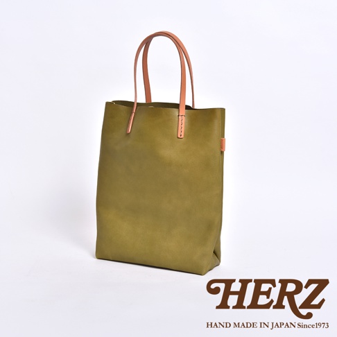 這是一款由日本HERZ職人手工製作的直式軟皮托特包，呈現橄欖綠色調，搭配天然原色的皮革提把，整體設計簡約俐落，展現出皮革自然的質地與細膩的縫線細節，圖片右下方印有HERZ品牌名稱與相關標語。