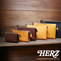 數個職人手作皮革三角底拉鍊手拿包，顏色包含深棕色、駝色及黑色，以不同尺寸並列展示於木質背景的層架上。