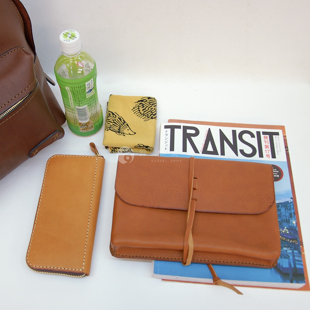 職人手作皮革休閒風後背包R-93，與一個棕褐色拉鍊皮夾、一個束帶的棕褐色皮革收納包、一本TRANSIT雜誌、一條黃色刺蝟圖案的布巾，以及一瓶綠色瓶身的飲品。