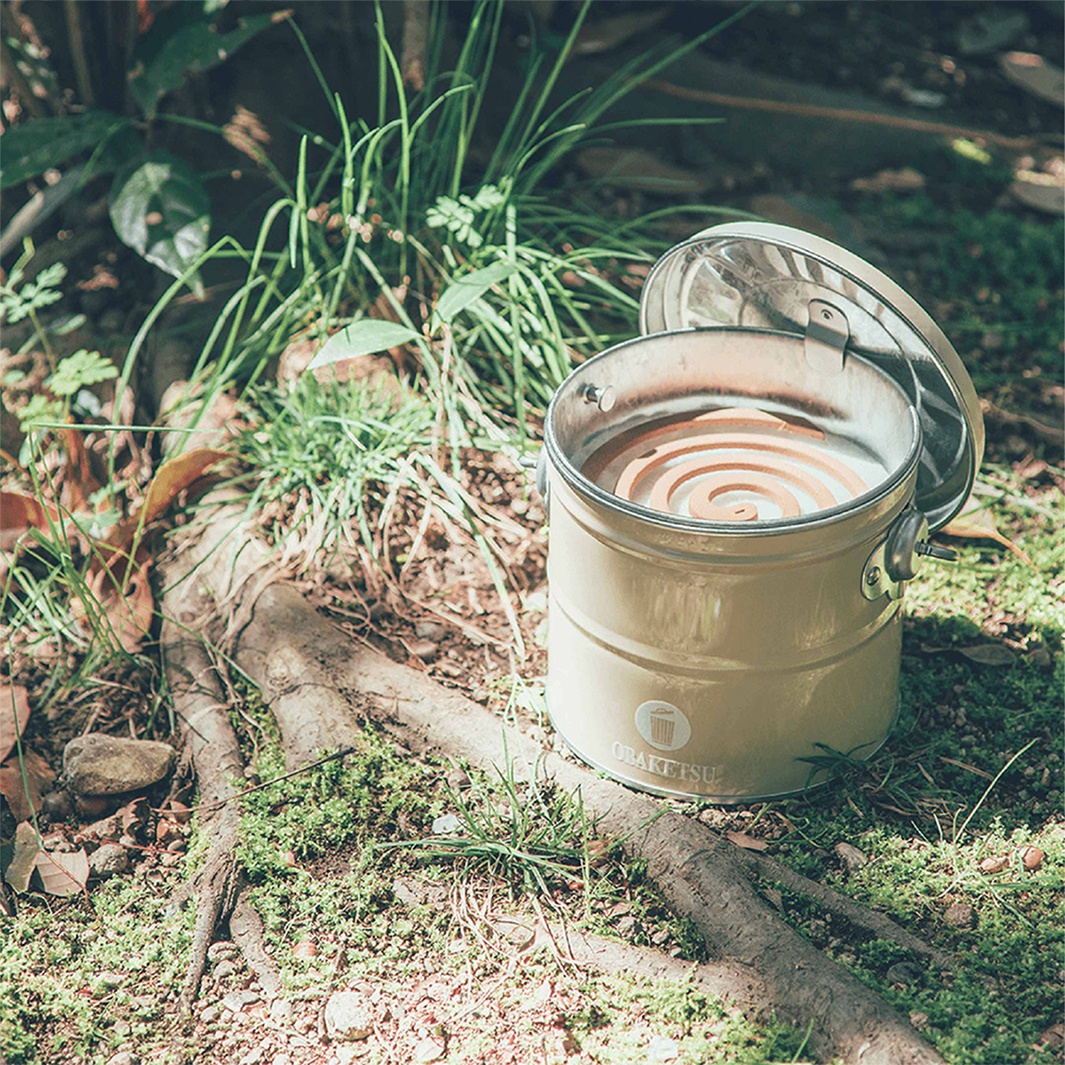 OBAKETSU 手工製蚊香桶，放置於充滿綠意與樹根的戶外草地上，香薰盤線條清晰可見，蓋子半開，展現其日製質感。