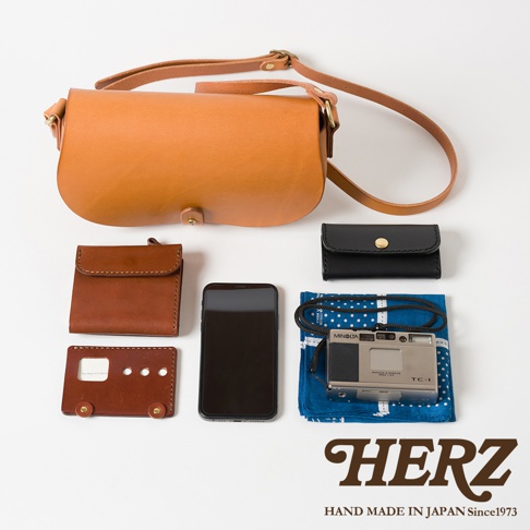 皮革迷你馬鞍包，附有可調式斜揹帶，搭配一個棕色皮革短夾、一個黑色皮革零錢包、一支手機、一台銀色復古相機和一塊藍色印花手帕，展現職人手作質感。