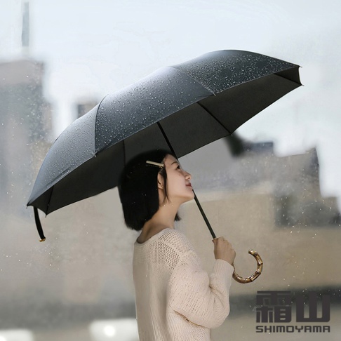 一位留著短髮的女性站在雨中，手持一把帶有精緻竹節手柄的黑色摺疊傘，雨滴落在傘面上，背景為模糊的城市建築，右下角印有霜山SHIMOYAMA品牌字樣。