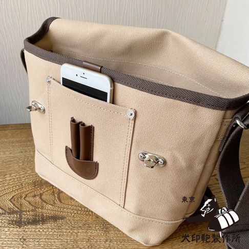 淺米色純棉帆布材質的郵差包，配有深棕色滾邊和背帶。包包正面有一個開放式口袋，內有一部白色手機，口袋上方有皮革拉環。另有兩個金屬鎖扣裝飾。