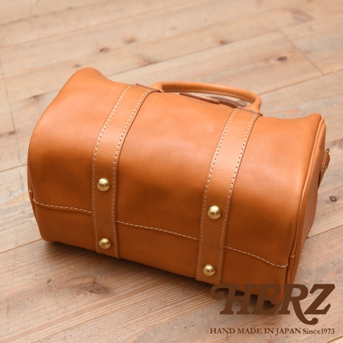 棕褐色皮革製成的圓筒形波士頓包，有兩條皮革提把，包身兩側各有一條皮革飾帶，飾帶上以黃銅色鉚釘固定，並以白色縫線裝飾。