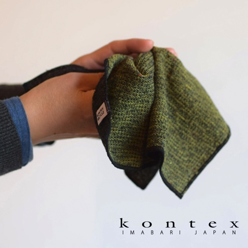 一塊綠色混織材質的長毛巾，邊緣為黑色，上面有白色標籤。毛巾被一隻手握著，背景為白色。