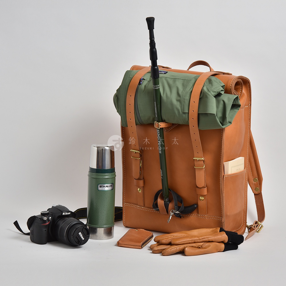 棕褐色皮革後背包，搭配綠色登山杖、綠色衣物、綠色保溫瓶、黑色相機及棕褐色皮手套。