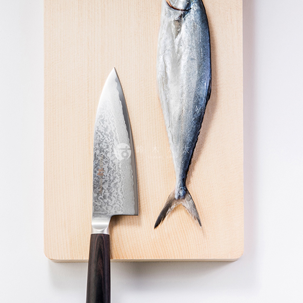 一把鋼製的 Deba 魚肉料理刀，刀身上有獨特的紋理，搭配深棕色刀柄，置於淺木色砧板上，旁邊有一條銀灰色的魚。