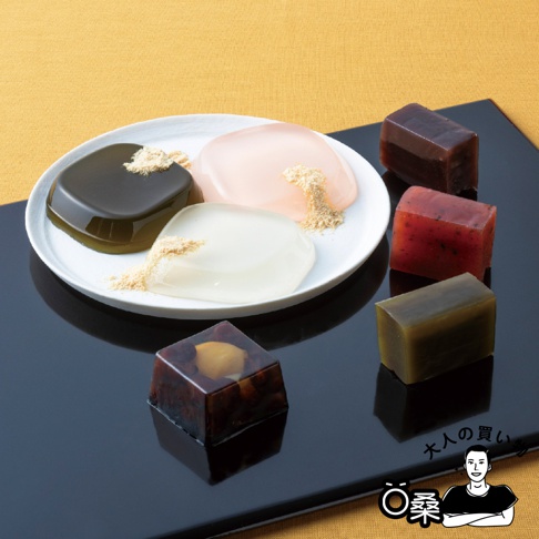 一張高品質的商品攝影照片，呈現了精緻的日式傳統甜點。盤中放置了三種不同顏色的透明果凍，表面撒上細緻的黃豆粉；盤旁擺放著數塊方形的羊羹，包含紅豆、栗子等多種口味。整體佈置在黑色的方形展示托盤上，背景為簡約的淺黃色織布紋理，右下角有一個人物圖案及大人的購物字樣。