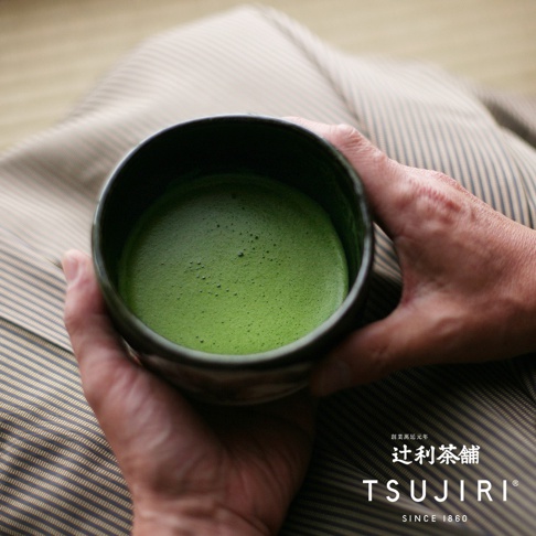 雙手捧著一個深色、粗糙質感的抹茶專用茶碗，碗內盛滿翠綠色、表面有細緻泡沫的抹茶。背景為帶有細條紋的布料。