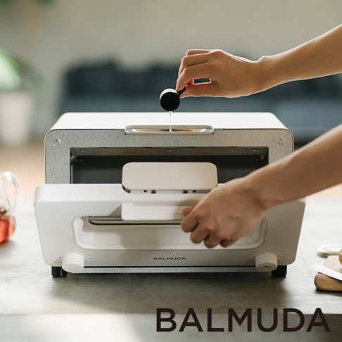 白色蒸氣烤麵包機，頂部有一個注水口，一隻手正在將水倒入。烤麵包機下方是打開的烤箱門，另一隻手正在將烤架推入。