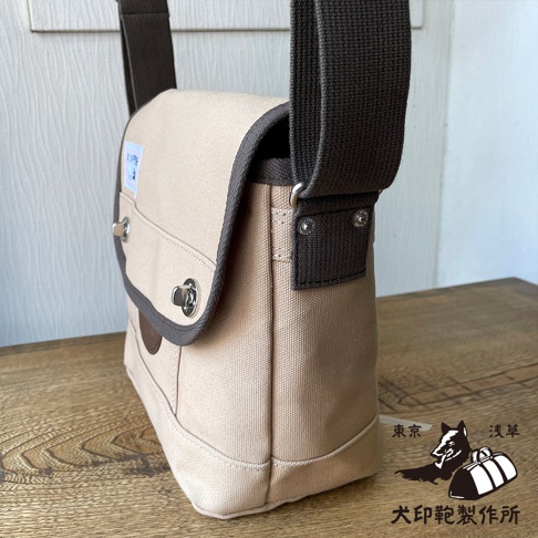 淺卡其色純棉帆布雙扣郵差包，搭配深棕色織帶和包邊，側面有品牌標誌。