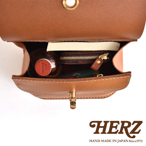 皮革翻蓋迷你後背包，展示內部空間，包含一瓶飲料、一個小錢包和一本筆記本，包款為棕褐色皮革材質，搭配金色鎖扣。