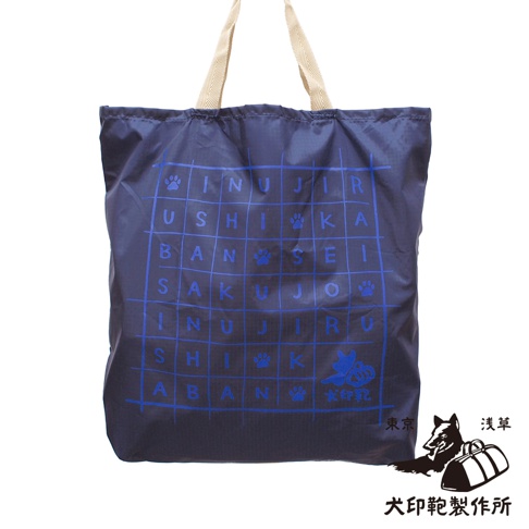 深藍色環保折疊收納購物袋，採用尼龍材質，正面印有藍色網格狀文字和狗爪圖案。袋子有兩個米白色織帶提把。