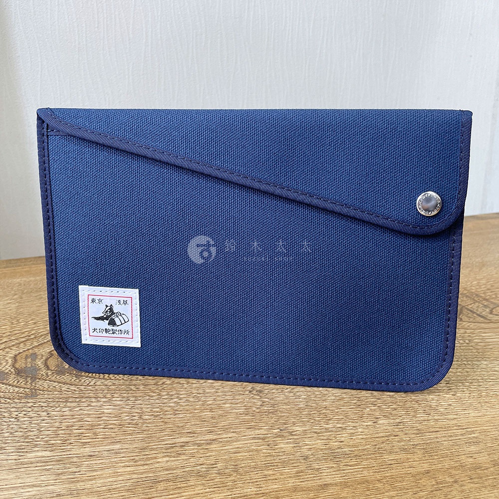 深藍色純綿帆布平板收納包，斜裁翻蓋設計，配有金屬按扣，左下角有品牌標誌貼紙。