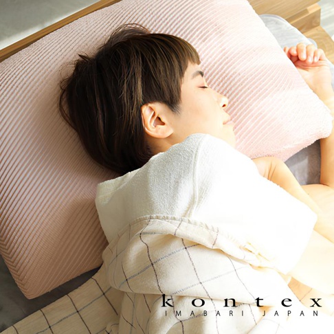 粉色今治萬用彈性枕套，表面有細緻的橫向壓紋，材質柔軟，搭配一名女子側臥，頭部枕於枕套上，身上蓋著一件格紋棉質蓋毯。