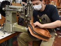 職人正在使用工業縫紉機縫製一個淺棕色皮革立體運動斜背包，包身具有粗獷的縫線及雙層拉鍊設計。