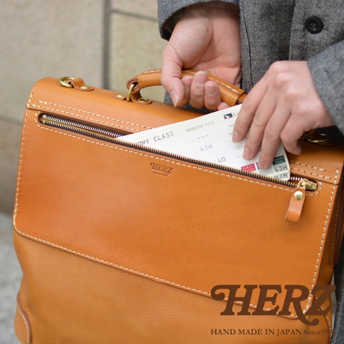 一款棕褐色皮革製作的波士頓包，包身結構清晰，縫線工整，配有金色拉鍊和金屬扣環。包包前方有一個拉鍊口袋，露出部分登機證。