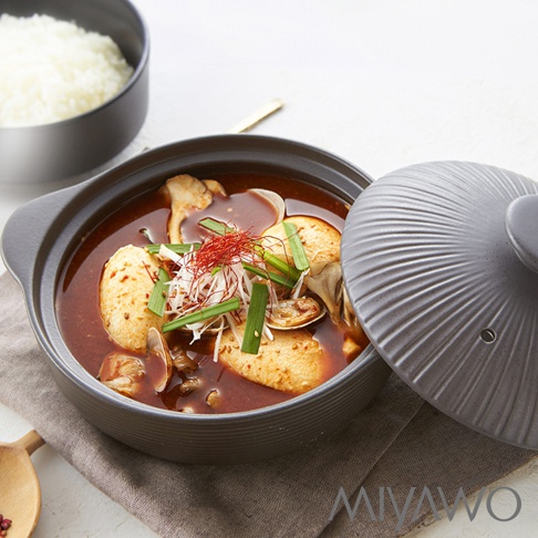 展示一個灰色的陶土湯鍋，鍋內盛有紅色的湯，湯中有豆腐、蛤蜊、香菇和蔥段，上方撒有辣椒絲。鍋蓋為灰色，帶有放射狀的紋路，部分蓋在鍋上。