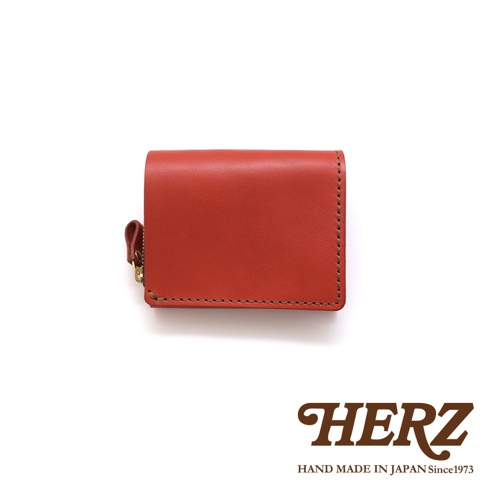 職人手作皮革小型雙折短夾，呈現橘紅色，採用真皮材質，縫線清晰，左側配有拉鍊與皮革拉環。