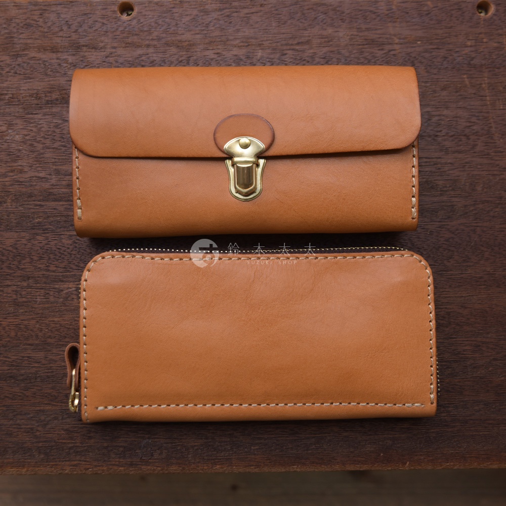 兩款淺棕色皮革長夾，上方為帶有金色金屬鎖扣的手風琴式長夾，下方為拉鍊式長夾，皆為手工縫製。