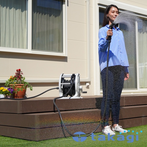 takagi 穩壓灑水組 Mercury 2 Twister 20m RT220CG7 婦女手持灑水器對著鏡頭微笑，背景是現代房屋的外部，有綠色的草坪和棕色的木質甲板。