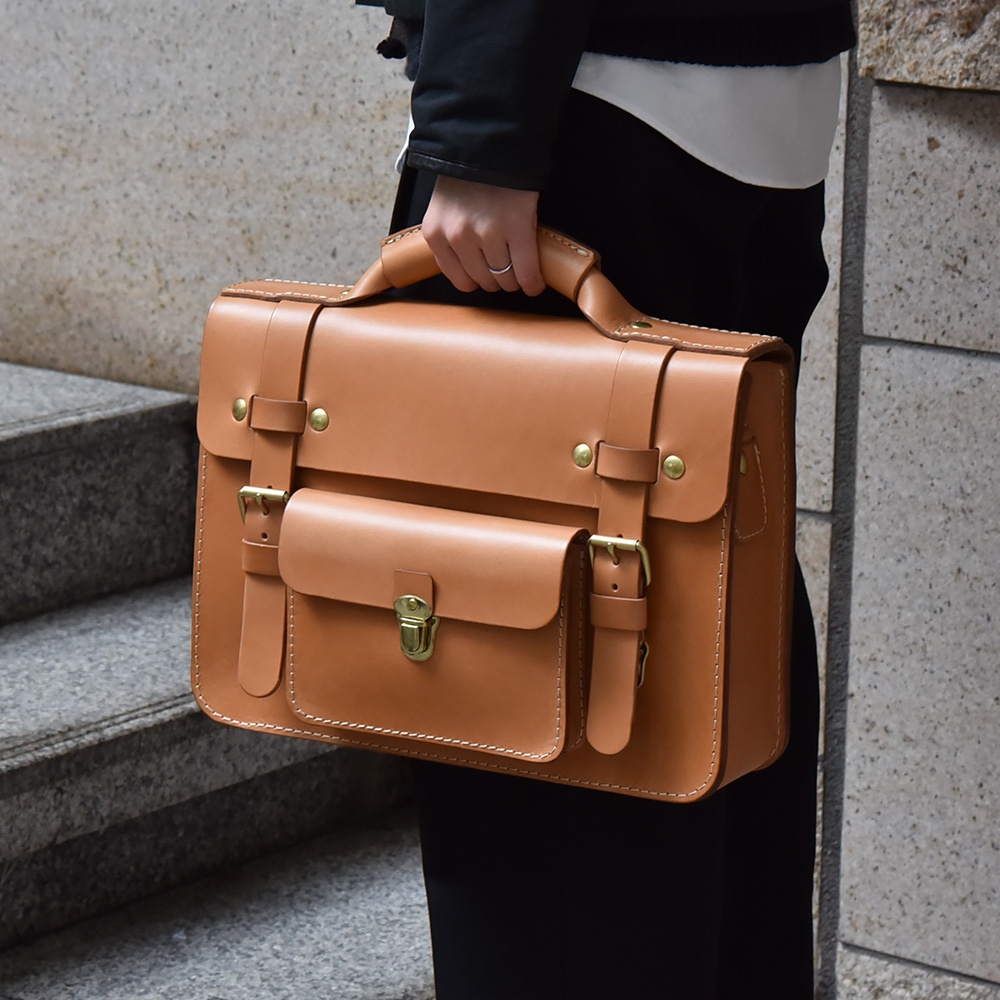 棕褐色皮革學院風兩用公事包，設計有翻蓋、兩個前袋、可調節扣帶及頂部提把，金屬扣件為復古黃銅色，縫線清晰。