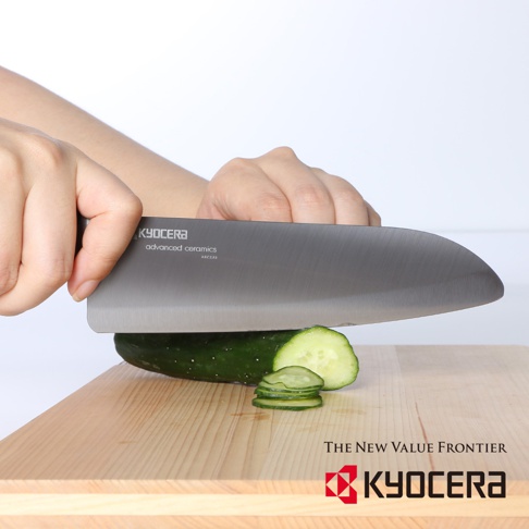黑刃精密陶瓷刀，刀身呈現金屬灰色，刀面上印有「KYOCERA advanced ceramics」字樣。刀鋒正在切一條綠色黃瓜，黃瓜已被切成數片薄片，置於淺木色砧板上。