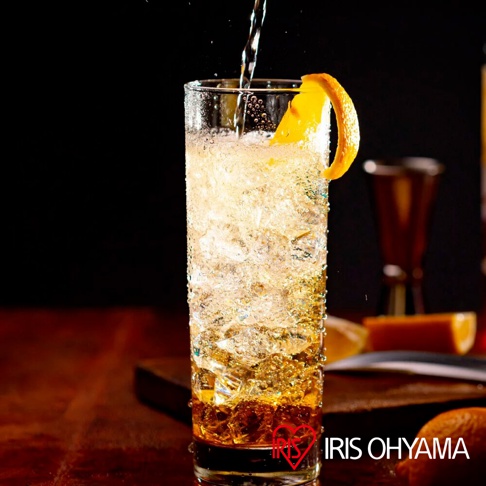 一個裝滿冰塊的透明玻璃杯，盛著琥珀色的氣泡飲品，頂部裝飾著一條橘色果皮，背景是深色，下方有「IRIS OHYAMA」的品牌標誌。