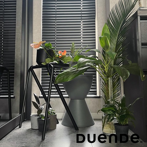 黑色金屬材質的低款吧檯椅，椅腳呈三腳架設計，上方平台放置有盆栽植物，旁邊擺放著深灰色花盆及其他綠植。