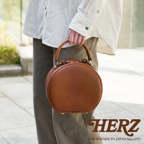 一名女子手持一個棕色圓形皮革側背包，包包上有HERZ品牌標誌和HAND MADE IN JAPAN Since1973字樣