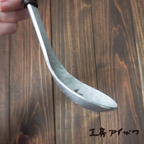 銀色鋁製湯勺，勺面有錘擊紋路，勺柄部分為深棕色材質，背景為木紋桌面。