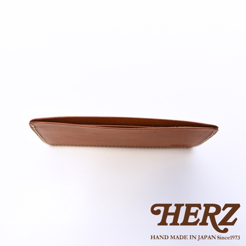從上方視角拍攝的HERZ棕色皮革紙鈔收納夾，皮夾呈現扁平修長的長方形結構，邊緣可見精緻的車縫細節，右下角印有品牌標誌與手工製作說明，整體風格簡約且質感厚實。