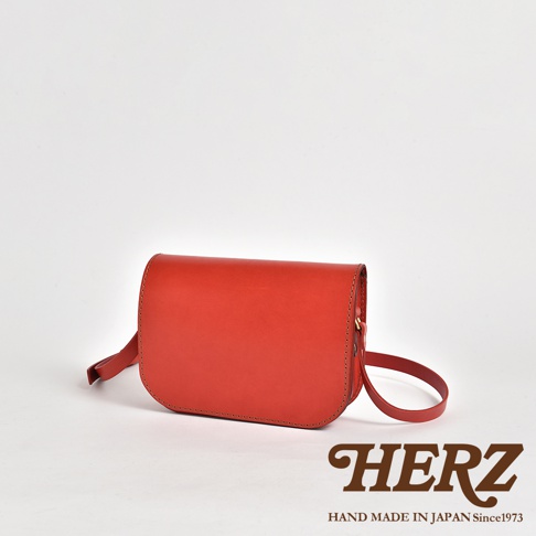 一個紅色的皮革翻蓋橫式單肩包，配有可調節的肩帶，包身線條簡潔，縫線清晰，呈現職人手作質感。