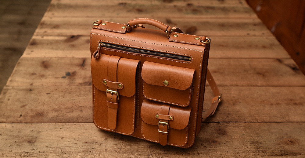棕色皮革製成的手工三用後背包，正面有三個帶有銅扣和皮帶的口袋，上方有一個拉鍊口袋和提把，背面可見部分肩帶，整體風格簡約實用。