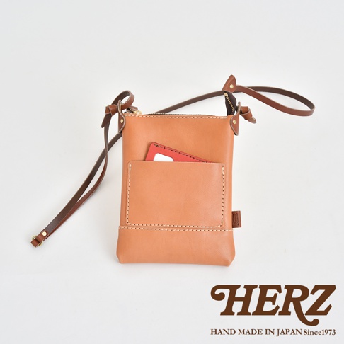 職人手作皮革直式隨身單肩包，淺棕色皮革，正面有一個較小的外袋，內有紅色卡片，搭配深棕色皮革背帶，整體簡約實用。
