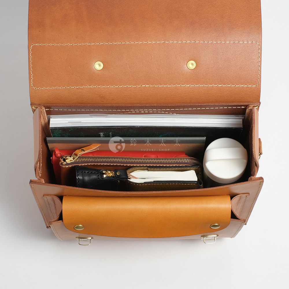 職人手作皮革橫式書包內部，裝有書籍、紅色皮夾、黑色筆記本和白色圓形小物。