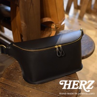 黑色皮革立體斜背包，配有金色雙拉鍊，簡約設計，展現職人手作質感。