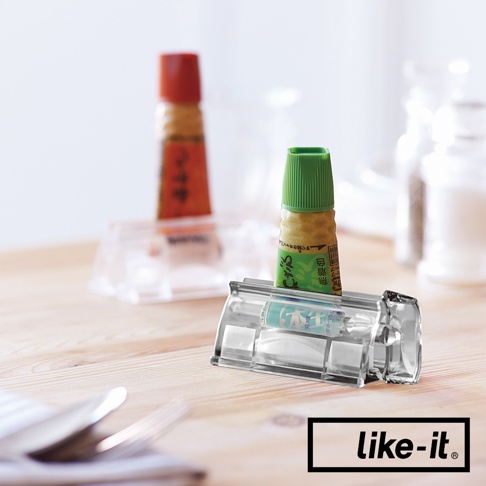 透明壓克力材質的手動轉轉擠牙膏夾，夾著綠色瓶蓋的調味料小瓶，背景有另一個紅色瓶蓋的調味料小瓶和鹽罐胡椒罐。