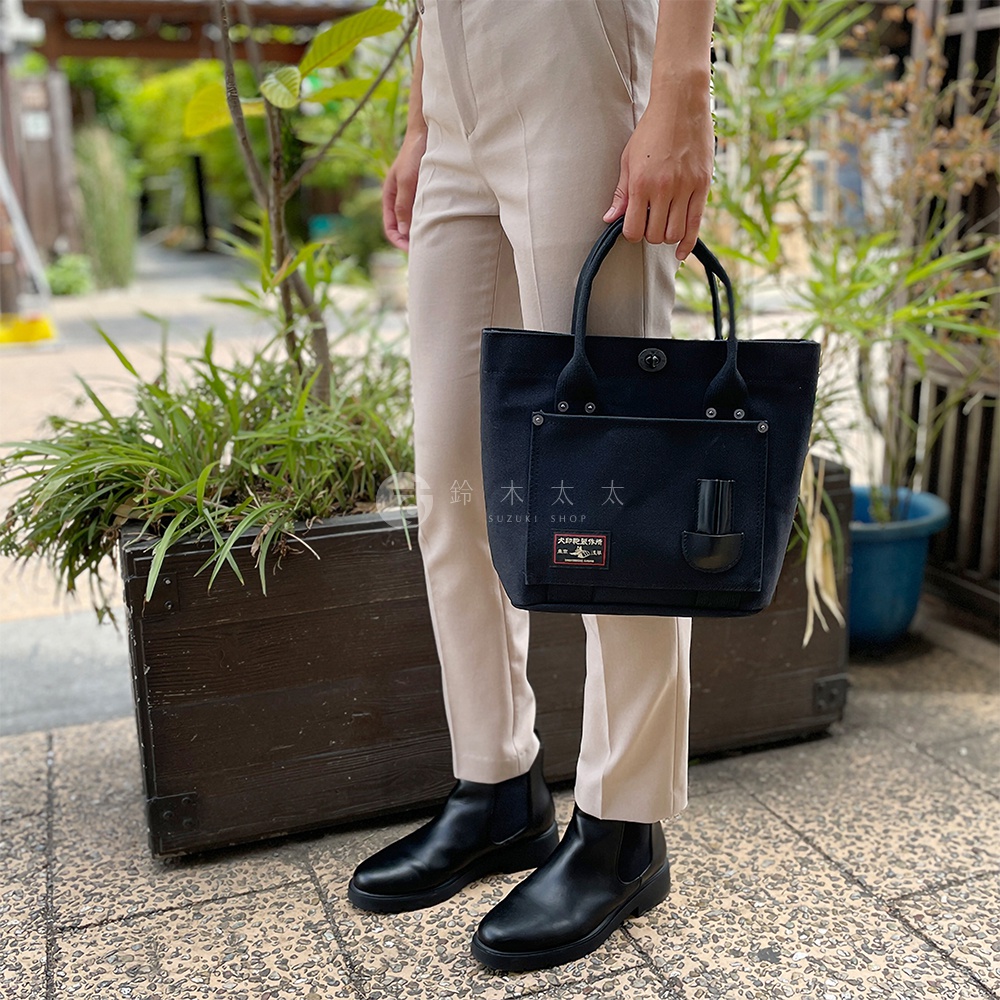 黑色帆布水桶托特包，附有兩個提把、一個外部口袋和一個小皮革掛環，包上還有一個品牌標籤。