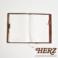 職人手作皮革手帳書套，打開的書頁內頁有方格，一條細繩連接兩頁，書套為棕色皮革，右下角印有HERZ LOGO。