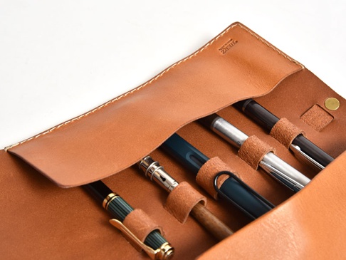 棕色皮革捲捲收納筆袋，內有六支不同顏色和材質的筆，筆袋邊緣有縫線，右側有金屬按扣。